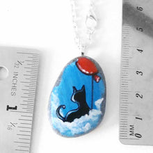 Load image into Gallery viewer, a small beach stone, hand painted with a little black cat, with a red balloon, sitting on clouds against a blue sky. available as a keepsake or pendant necklace
