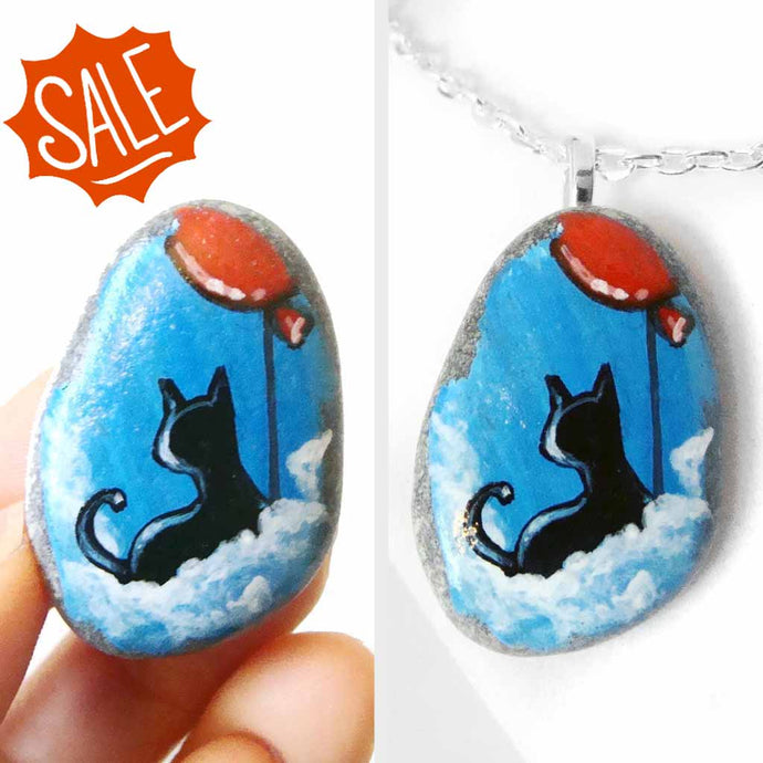 a small beach stone, hand painted with a little black cat, with a red balloon, sitting on clouds against a blue sky. available as a keepsake or pendant necklace
