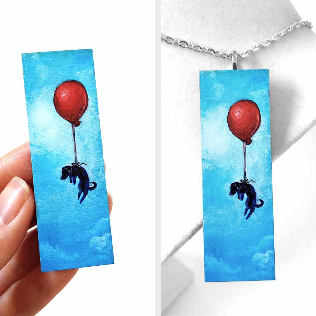 a wood rectangle hand painted with art of a black dachshund dog, with a red balloon, floating through a blue sky. available as a keepsake or pendant necklace