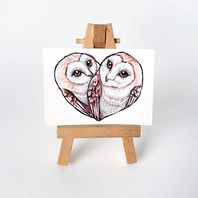 An ACEO original painting, placed on an easel, featuring two barn owls together, forming the shape of a heart