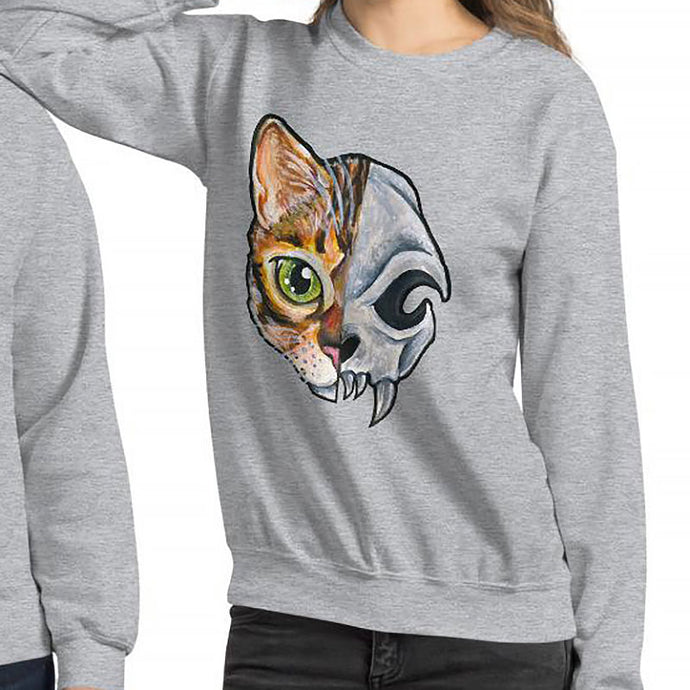 A woman is wearing a unisex sweatshirt in the colour sport grey, printed with art split into two: the left side features the face of a bengal cat, and the right side features an evil looking cat skull.