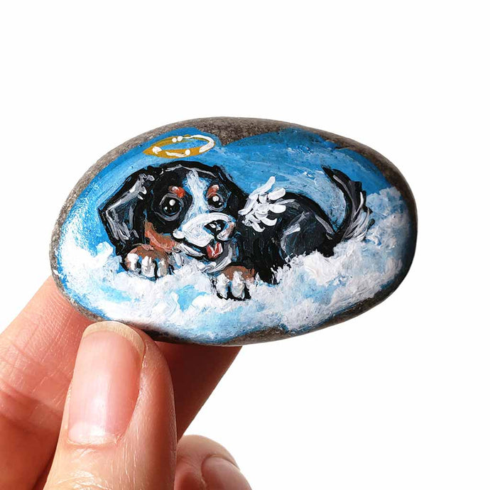 a smooth beach stone, hand painted with art of a bernese mountain dog, as an angel int he clouds
