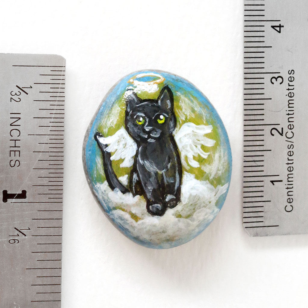 Rock art of a black cat as an angel, painted on a small beach stone, next to two rulers to show its size: 1 5/16
