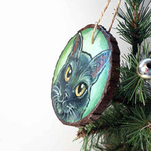 Load image into Gallery viewer, a round wood ornament, hand painted with a portrrait of a black cat&#39;s face, with yellow eyes, against a soft green background
