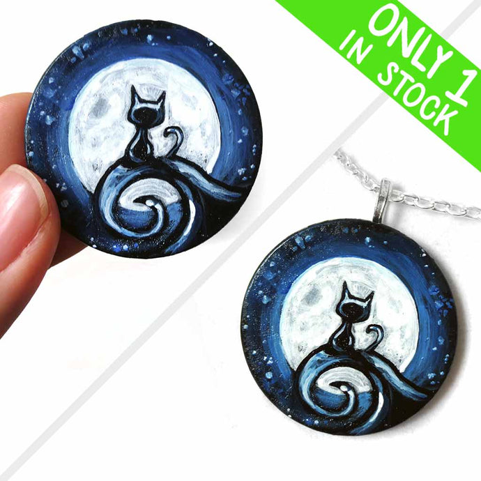 a wood disc, hand painted with a small black cat sitting on a curly hill, against a large full moon and starry night sky. this piece is available as a keepsake o rpendant necklace