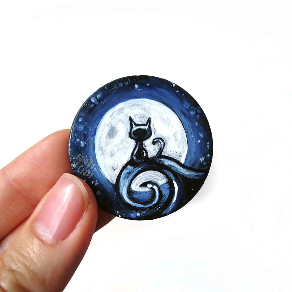a wood disc, hand painted with a small black cat sitting on a curly hill, against a large full moon and starry night sky. this piece is available as a keepsake o rpendant necklace