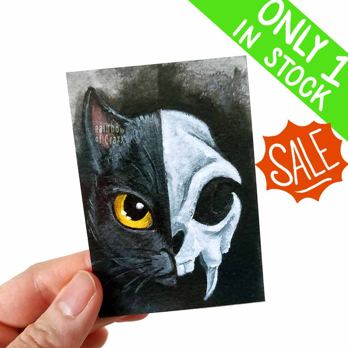 an aceo art print features two sides of a black cat: a cute kitty on the one side and a creepy cat skull on the other.