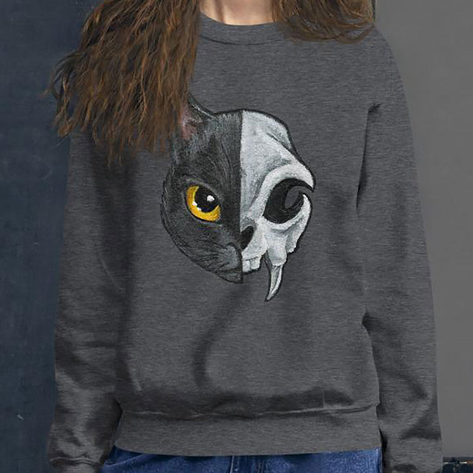 A woman is wearing a unisex sweatshirt in the colour dark heather grey, printed with art of a split image: the left side features a yellow eyed black cat, and the right side features an evil looking cat skull