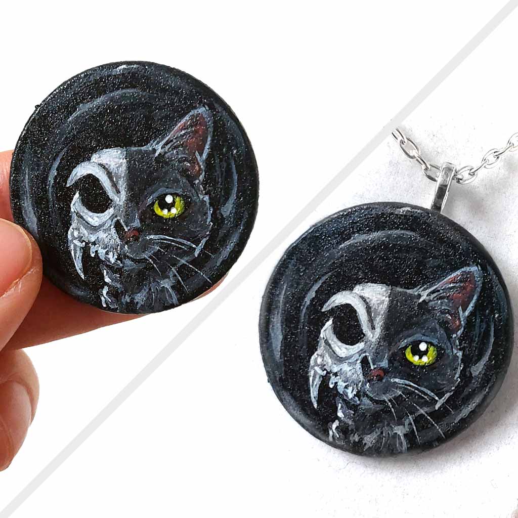 a circle shaped wood disc, hand painted with a split image: a black cat's face on the right side, and a cat skull on the left side. available as a keepsake or pendant necklace