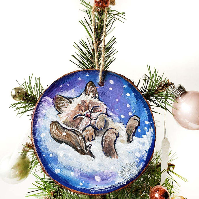 A Christmas tree wood ornament, hand painted with art of a Himalayan cat sleeping on snow.