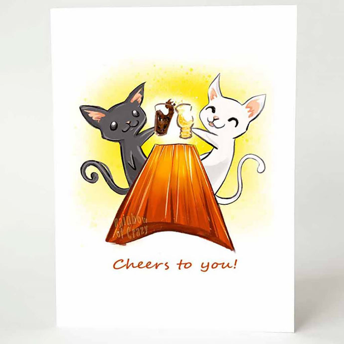 a greeting card, with art of a black cat and white cat toasting each other's beer glasses while sitting at a large wooden table. the card reads, 'cheers to you!'
