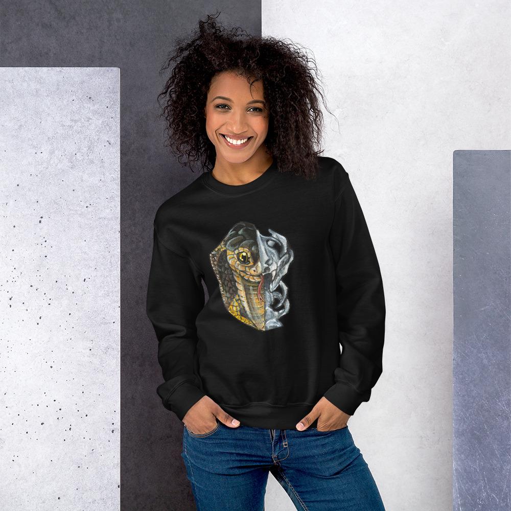 A woman is wearing a unisex sweatshirt in the colour black, printed with an illustration split into two: the left side features the face of a cobra snake, and the right side features an evil looking snake skull.