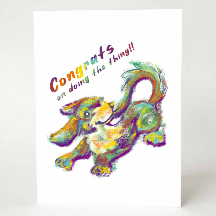a greeting card featuring a dog catching her own tail, painted with rainbow colours. above reads, congrats on doing the thing!!