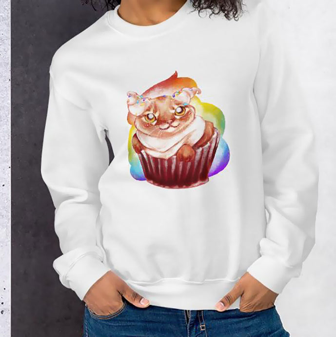 A woman wears a unisex sweatshirt in the colour white, featuring art of a cat painted as a red velvet cupcake with rainbow sprinkles.