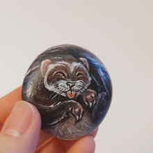 Load and play video in Gallery viewer, a video of a hand holding a small beach stone, hand painted with a portrait of a smiling ferret with its tongue out
