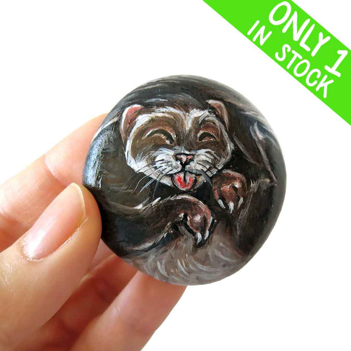 a small beach stone, hand painted with a portrait of a smiling ferret with its tongue out