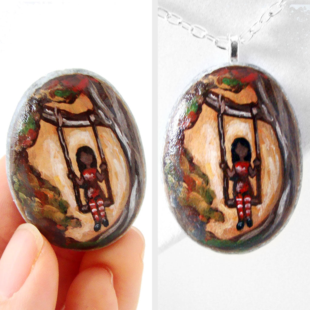 small rock art: a beach stone is hand painted with a girl in red, sitting on a tree swing, available as either a keepsake or a necklace