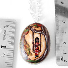 Load image into Gallery viewer, a beach rock with art of a woman in red, sitting on a tree swing. available as a necklace, the stone is next to two rulers to show its size: 1 1/4&quot; x 1 1/8&quot; or 3.1 cm x 2.8 cm
