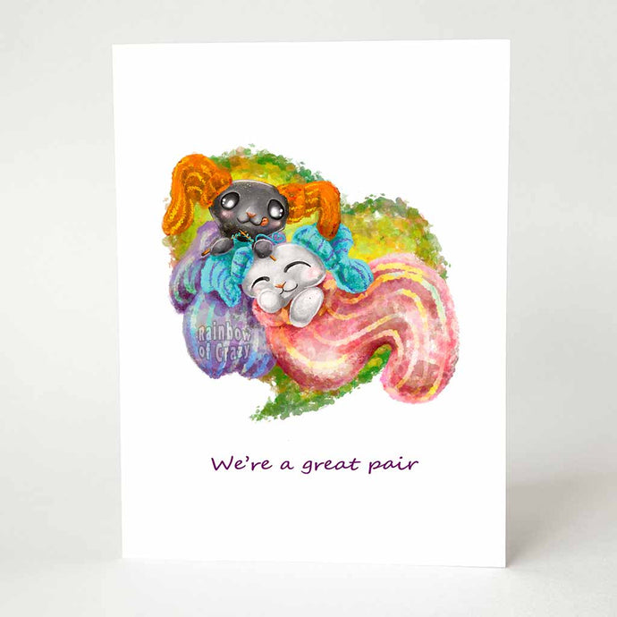 a greeting card featuring art of a black cat and white cat cuddling together in giant socks with smaller socks on their ears. The black cat finishes knitting one of the white cat's socks. the card reads, we're a great pair.