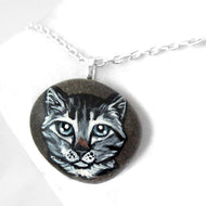 a small beach rock featuring a painting of a grey tabby cat with blue / grey eyes, available as either a keepsake, fridge magnet, or a pendant necklace