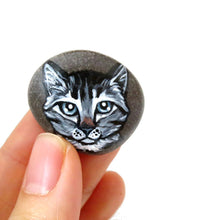 Load image into Gallery viewer, a small beach rock featuring a painting of a grey tabby cat with blue / grey eyes, available as either a keepsake or a pendant necklace
