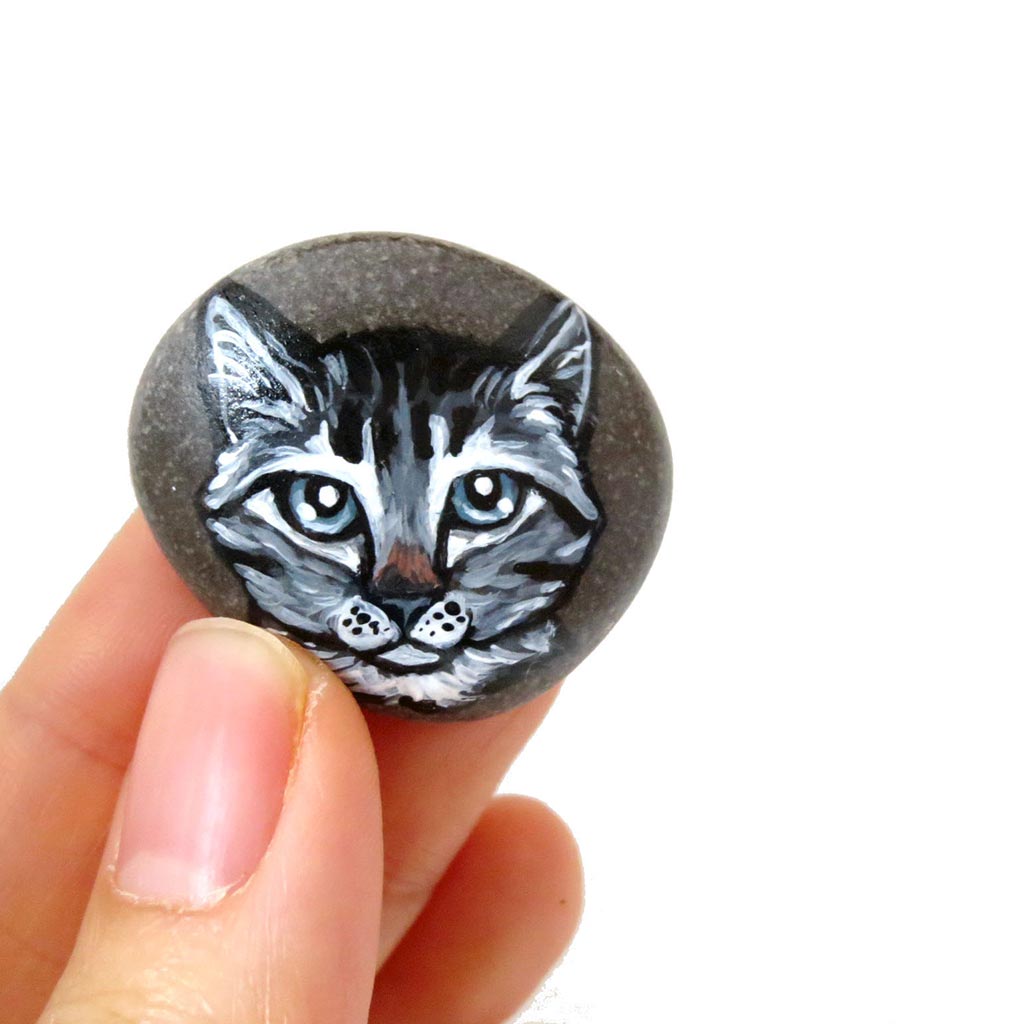 a small beach rock featuring a painting of a grey tabby cat with blue / grey eyes, available as either a keepsake or a pendant necklace