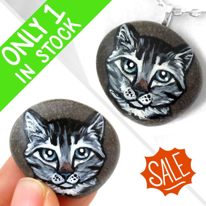 a small beach rock featuring a painting of a grey tabby cat with blue / grey eyes, available as either a keepsake or a pendant necklace