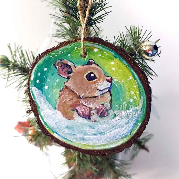 a christmas wood ornament, hand painted with the portrait of a syrian hamster, standing on the snow, watching snowflakes fall against a green and yellow sky