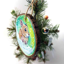 Load image into Gallery viewer, a christmas wood ornament, hand painted with the portrait of a syrian hamster, standing on the snow, watching snowflakes fall against a green and yellow sky
