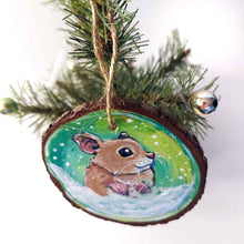 Load image into Gallery viewer, a christmas wood ornament, hand painted with the portrait of a syrian hamster, standing on the snow, watching snowflakes fall against a green and yellow sky
