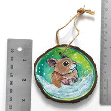 Load image into Gallery viewer, a christmas wood ornament, hand painted with the portrait of a syrian hamster, standing on the snow, watching snowflakes fall against a green and yellow sky
