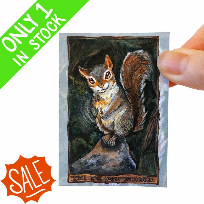 An aceo art print of the happy squirrel tarot card, from the animism tarot: a mischievous looking squirrel sits on a rock in the forest, a lit match in hand.