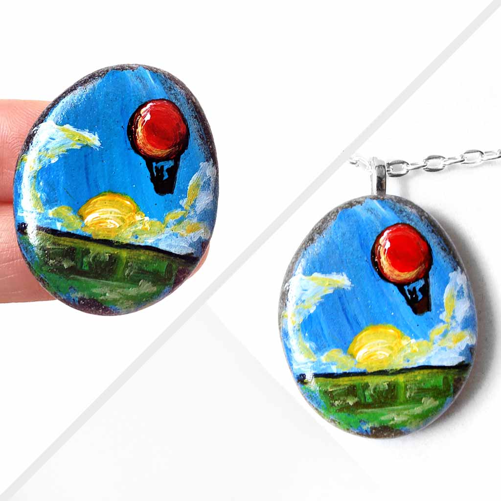 a small rock art of a red hot air balloon, floatiing in the sky over a green landscape, sun shining. available as a keepsake or pendant necklace