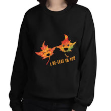 Load image into Gallery viewer, A woman is wearing a unisex sweatshirt in the colour black, featuring art of two smiling maple leaves with the words, &quot;I Be-Leaf In You&quot;
