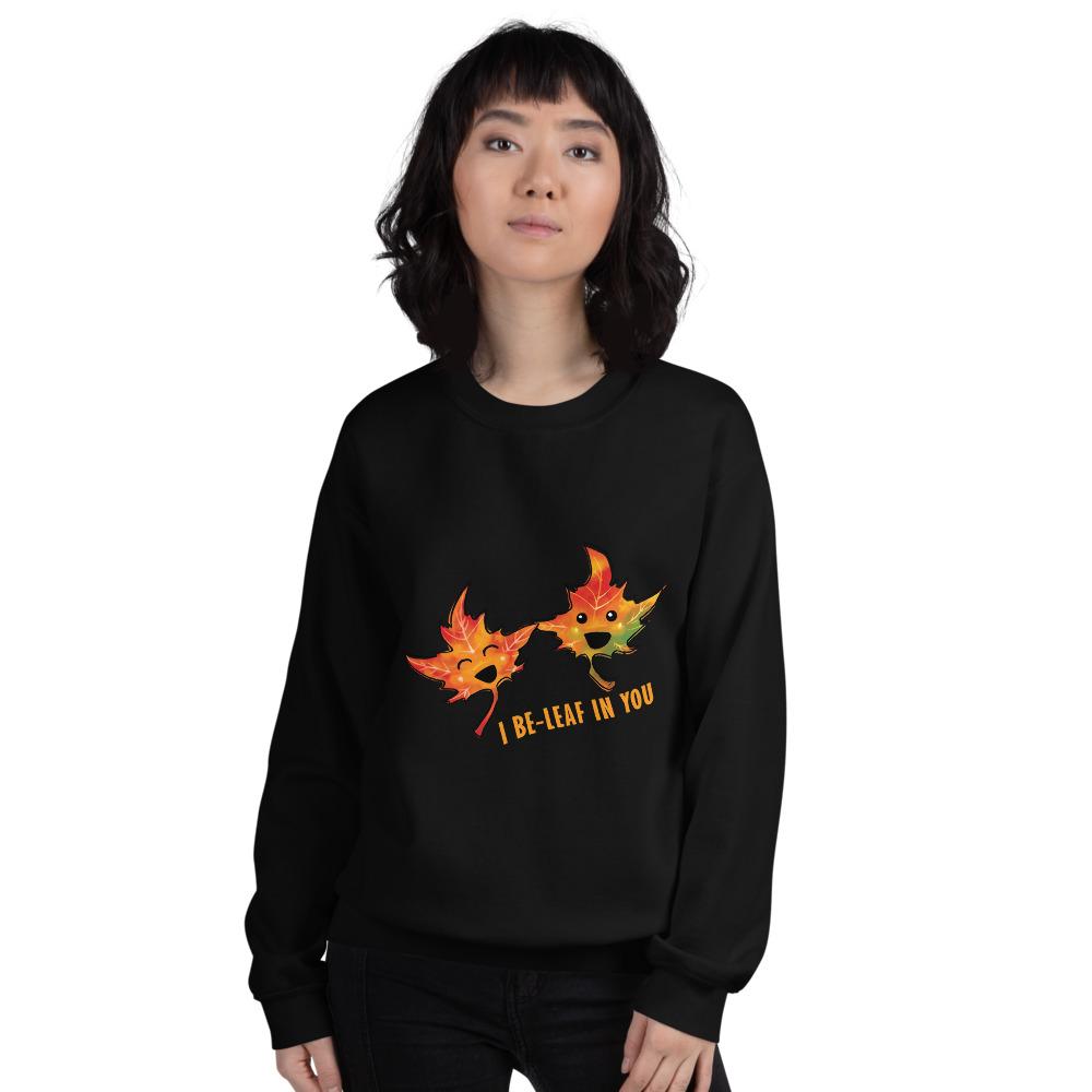 A woman is wearing a unisex sweatshirt in the colour black, featuring art of two smiling maple leaves with the words, 