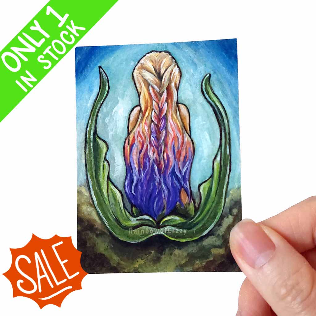 an aceo art print features a mermaid, from the back, with soft multi colour hair in blonde, pink and purple.