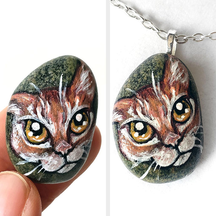 a small beach rock painted with of an orange tabby cat with golden yellow eyes, available as either a keepsake or a pendant necklace