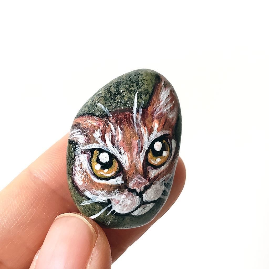 a small beach rock painted with of an orange tabby cat with golden yellow eyes, available as either a keepsake or a pendant necklace