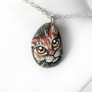 a small beach rock painted with of an orange tabby cat with golden yellow eyes, available as either a keepsake or a pendant necklace