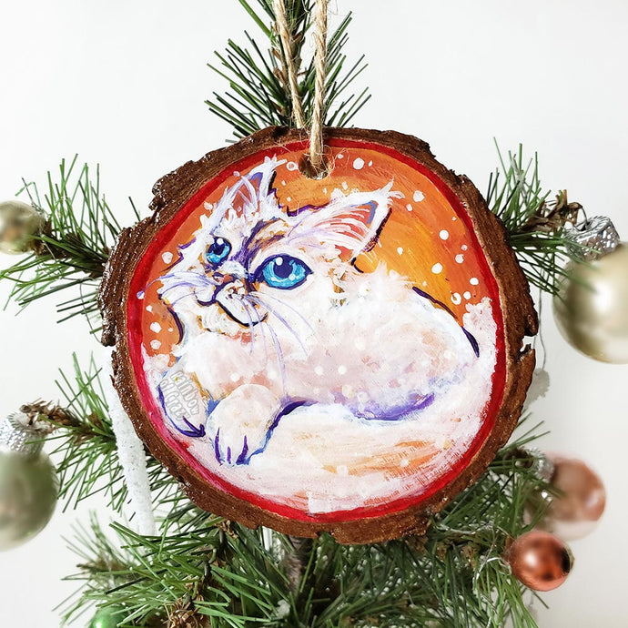 A wooden ornament with art of a white Ragamuffin cat with blue eyes, watching the snow fall