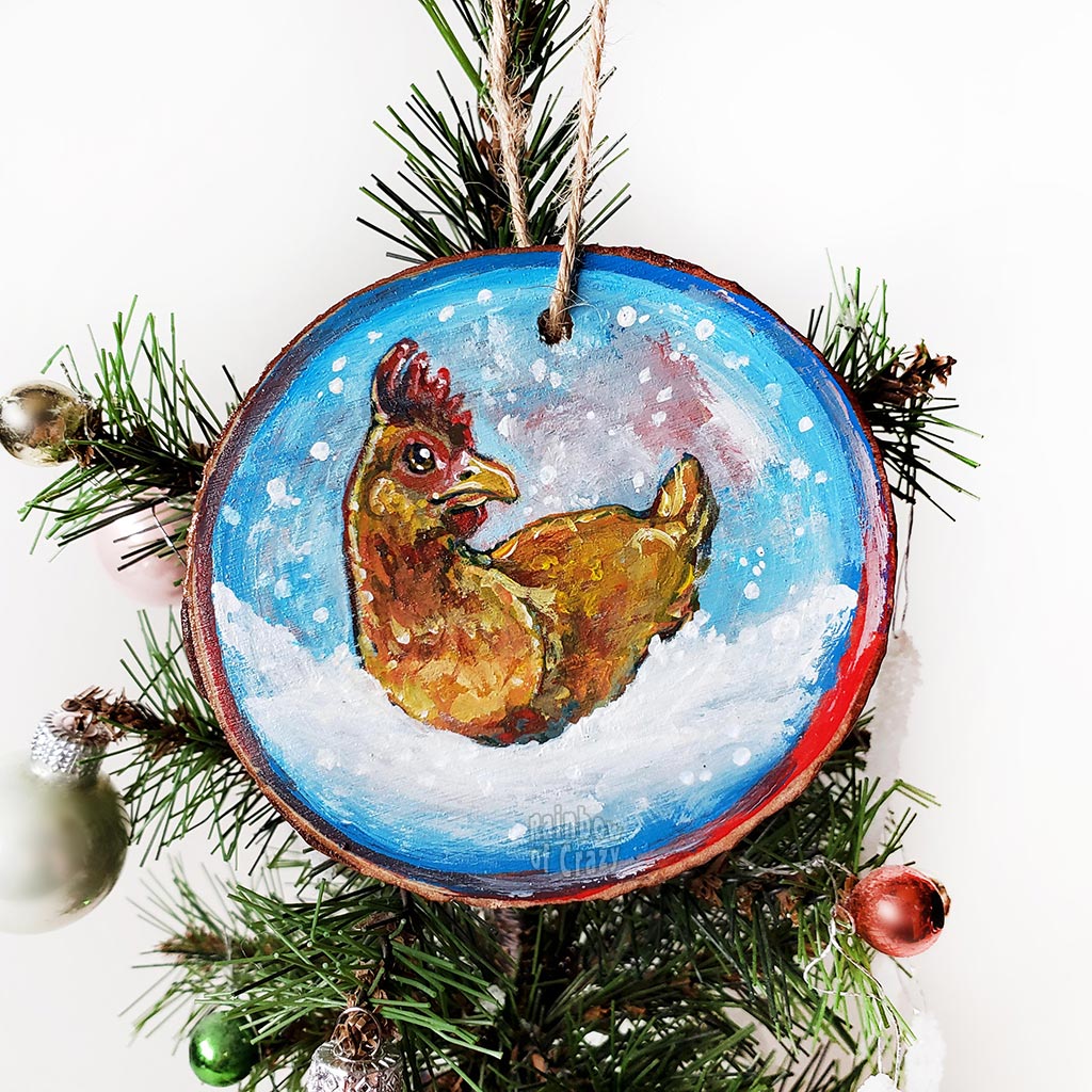A Christmas ornament with a painting of a chicken, sitting on snow as snowflakes fall. The ornament is painted in blues and reds.