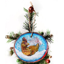 Load image into Gallery viewer, A holiday ornament with art of a chicken, sitting on snow as snowflakes fall. The ornament is painted in blues and reds.
