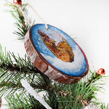 Load image into Gallery viewer, A holiday ornament with art of a chicken, sitting on snow as snowflakes fall. The ornament is painted in blues and reds.
