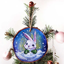 Load image into Gallery viewer, A wood Christmas ornament features art of a white rabbit, peeking out from behind the snow, wearing green mittens.
