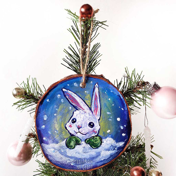 A wood Christmas ornament features art of a white rabbit, peeking out from behind the snow, wearing green mittens.
