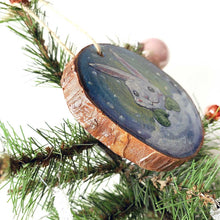Load image into Gallery viewer, A Christmas ornament, painted on a wood slice, features art of a white rabbit, peeking out from behind the snow, wearing green mittens.
