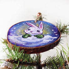 Load image into Gallery viewer, A holiday ornament, painted on a wood slice, features art of a white rabbit, peeking out from behind the snow, wearing green mittens.
