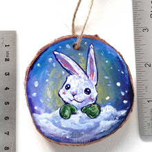 Load image into Gallery viewer, A Christmas ornament with a painting of a white rabbit with green mittens, painted on a wood slice, is displayed next to two rulers to show its size: 2 15/16 x 2 15/16 inches or 7.5 x 7.5 cm,
