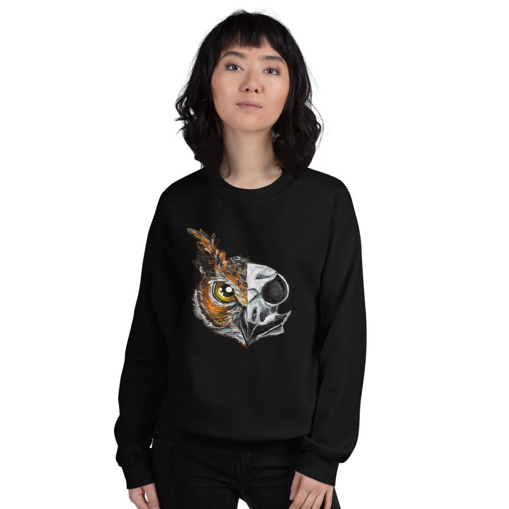 A woman wears a unisex sweatshirt in the colour black, printed with an illustration of a split image: the left side features an owl's face, and the right side features an evil looking owl skull.