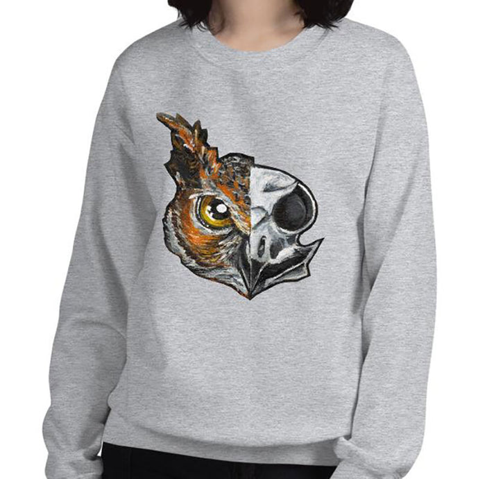 A woman wears a unisex sweatshirt in the colour sport grey, printed with artwork of a split image: the left side features an owl's face, and the right side features an evil looking owl skull.